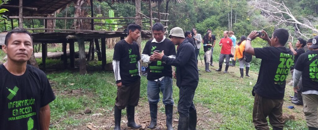 Five indigenous men wearing matching black shirts stand in front of a wood hut, looking at their phones.