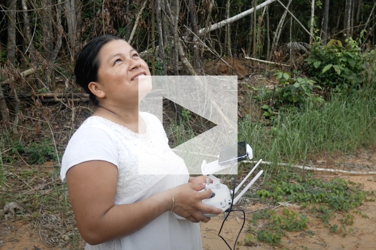 A young indigenous woman holds a drone controller in her hands. She looks into the sky, surrounded by rainforest.