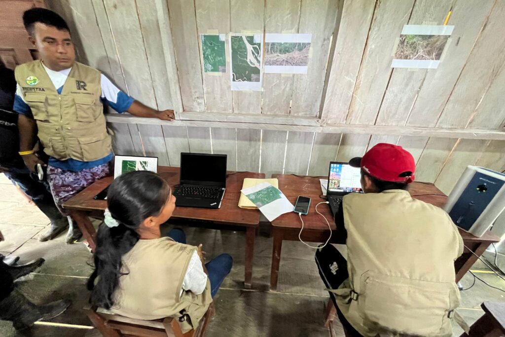 RFUS forest monitors in Loreto, the Peruvian Amazon, examine satellite images on laptops to track deforestation threats. The scene depicts a blend of traditional settings and advanced technology, with satellite maps and electronic devices on a wooden table.