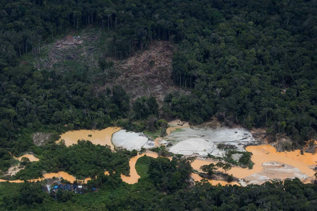 The image shows a deforested area in the midst of a lush rainforest, indicative of illegal mining activities. There are pools of water contaminated with mining residue, and makeshift structures are visible, contrasting sharply with the surrounding untouched forest. This is a representation of the environmental impact caused by such activities in the Yanomami Indigenous Land.