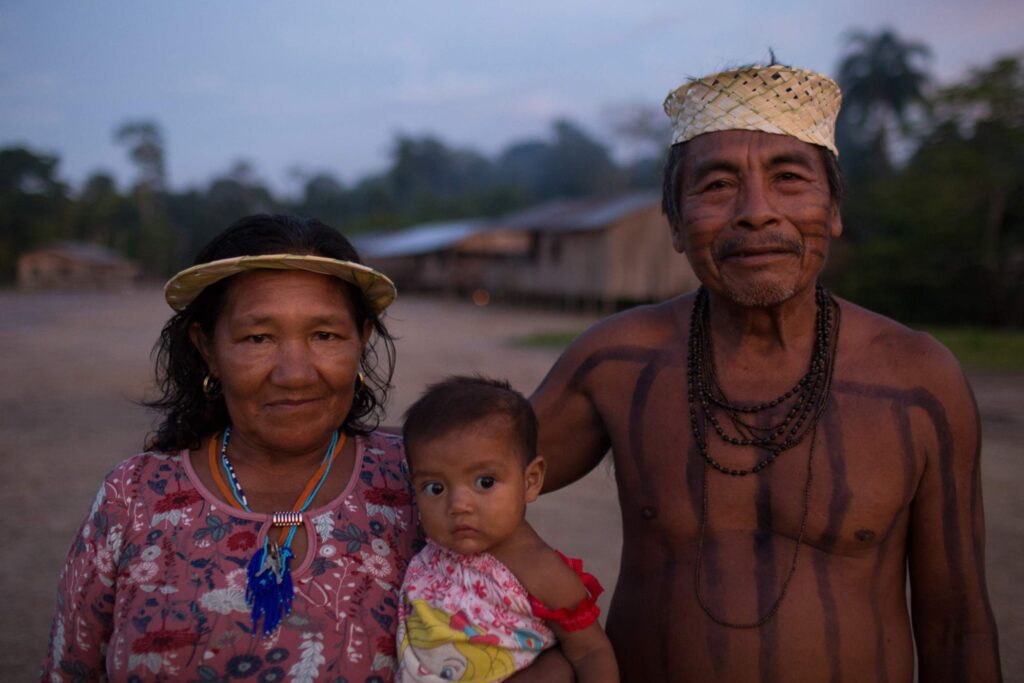 A portrait of a Kanamari family from the Massapê village, in the Indigenous Territory of Vale do Javari, Amazonas. The image features Eduardo Kanamari, also known as Dyanim in the Kanamari language, wearing a traditional woven hat and body paint, alongside his wife Zefinha, who is adorned with a distinctive necklace and is holding a young child.