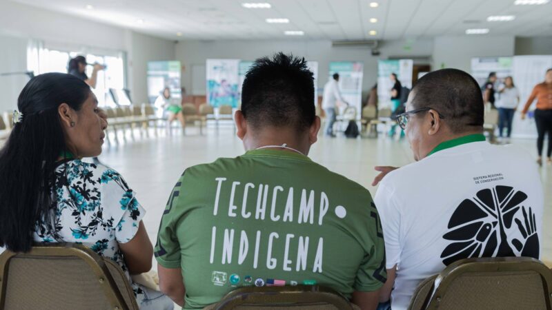 Three indigenous individuals seated with their backs to the camera. The shirt of the person in the center reads: Indigenous Tech Camp.